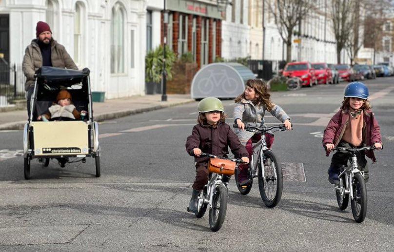 A father rides a Christiania tricycle with a baby on board while three primary school–aged children cycle with him.